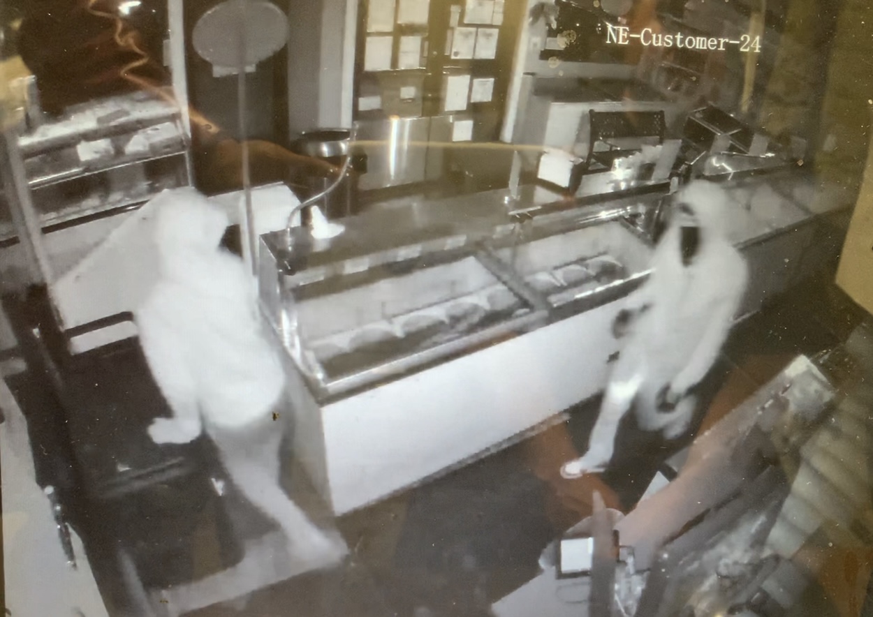 Surveillance footage of two individuals in hooded attire inside a dark, seemingly closed store. One person is near a counter with glass display cases, the other is to the right, both partially obscured.