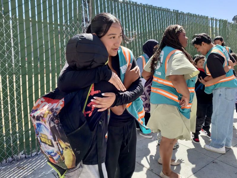 People gather near a chain-link fence. A child wearing a colorful backpack hugs an adult. Some people wear safety vests and appear involved in an organized activity or event.