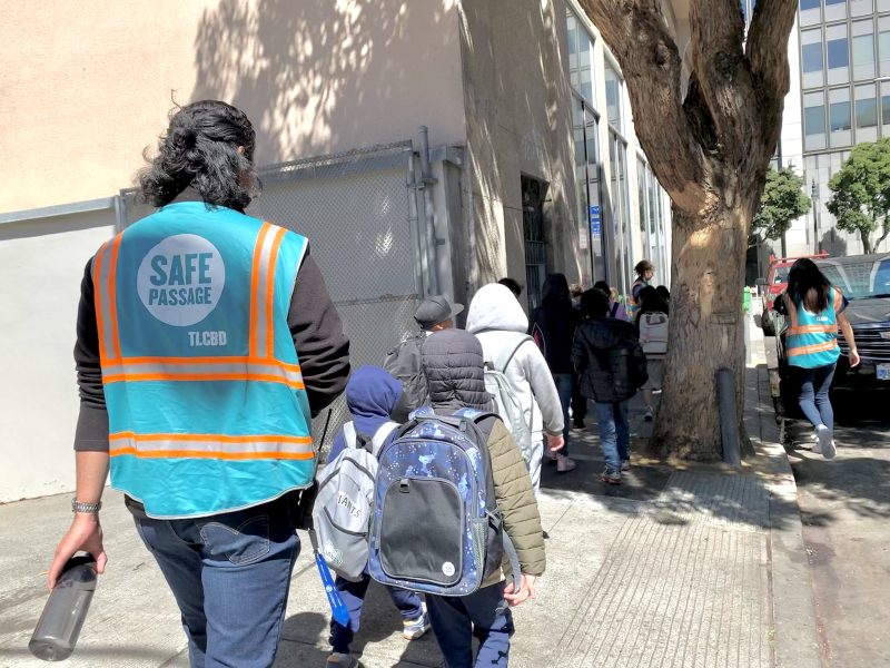 A group of people, including children with backpacks, walk down a sidewalk. Two adults wear bright blue vests labeled "SAFE PASSAGE." They are supervising and accompanying the group.