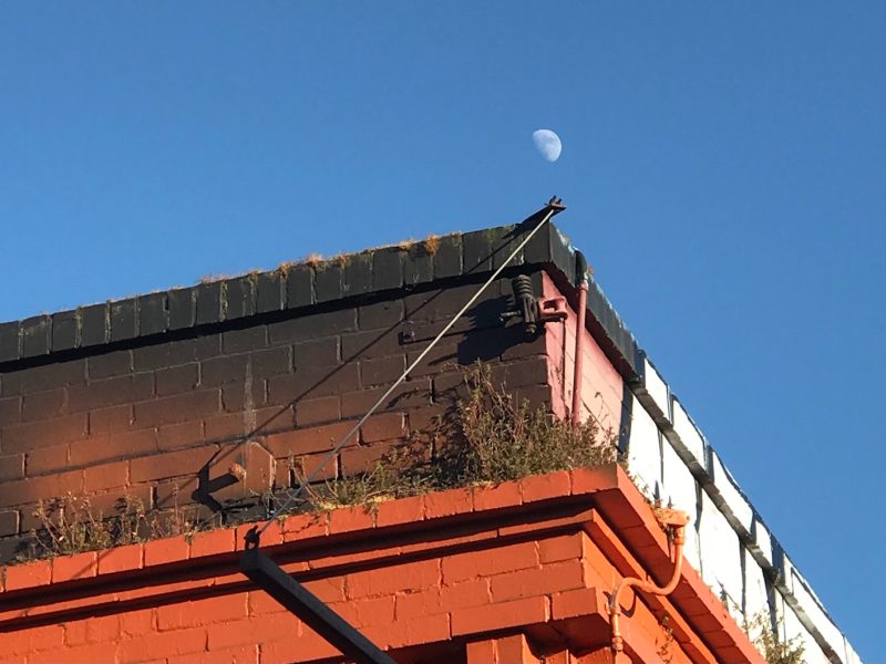 A partially visible moon against a blue sky, seen above the flat roof of a multicolored brick building with visible cables and some vegetation growth on the roof.