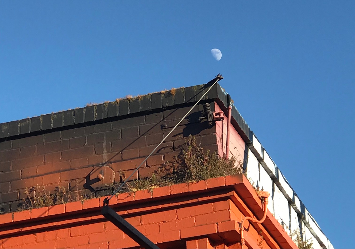 A partially visible moon against a blue sky, seen above the flat roof of a multicolored brick building with visible cables and some vegetation growth on the roof.