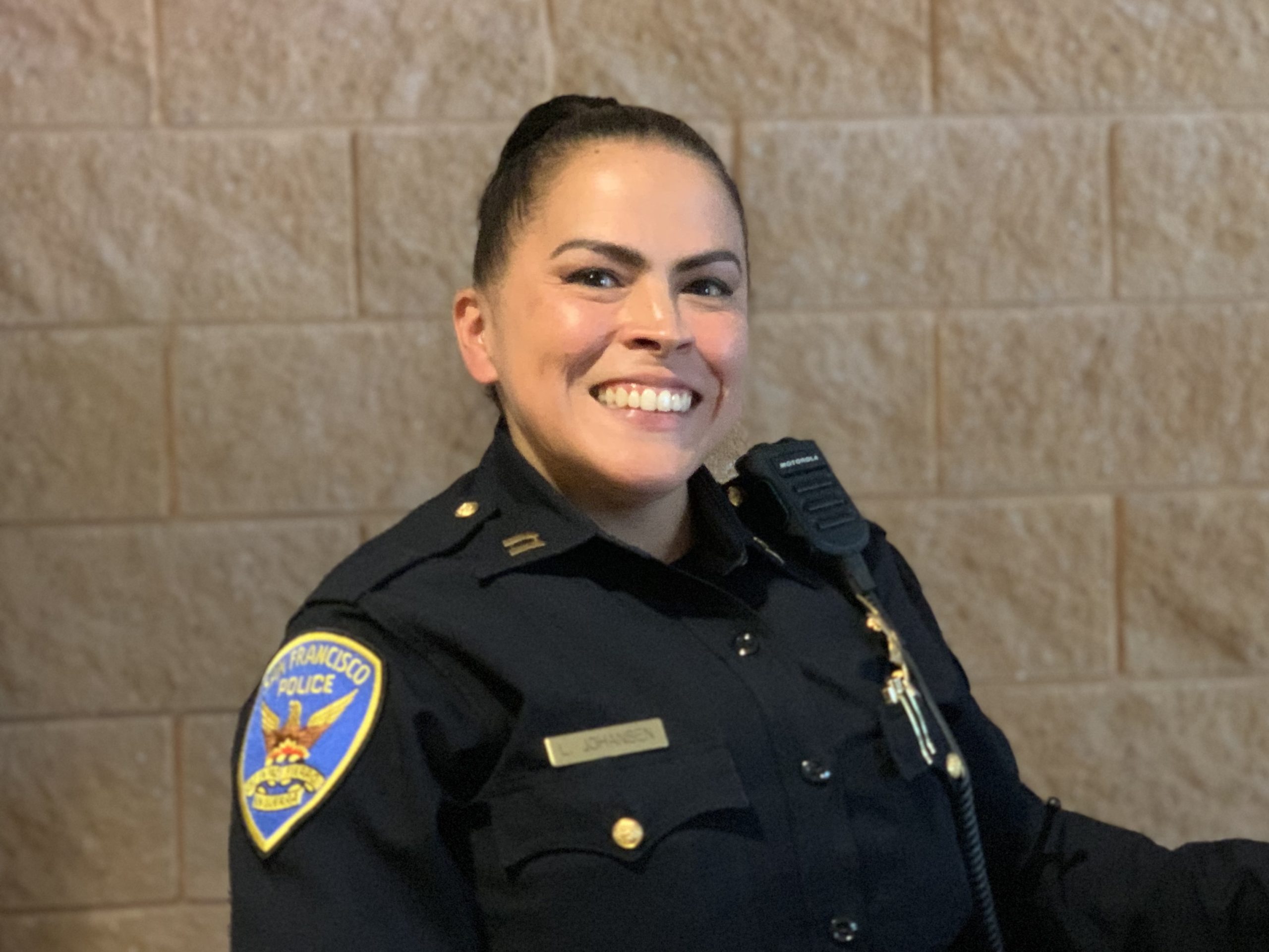 A police officer in uniform smiles while standing in front of a brown brick wall.