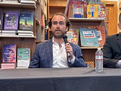 A man with curly hair and a beard, wearing a blue blazer, speaks into a microphone at a table in front of a bookshelf filled with books. A water bottle is on the table.