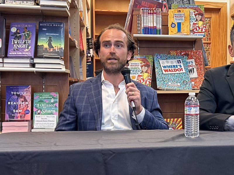 A man with curly hair and a beard, wearing a blue blazer, speaks into a microphone at a table in front of a bookshelf filled with books. A water bottle is on the table.