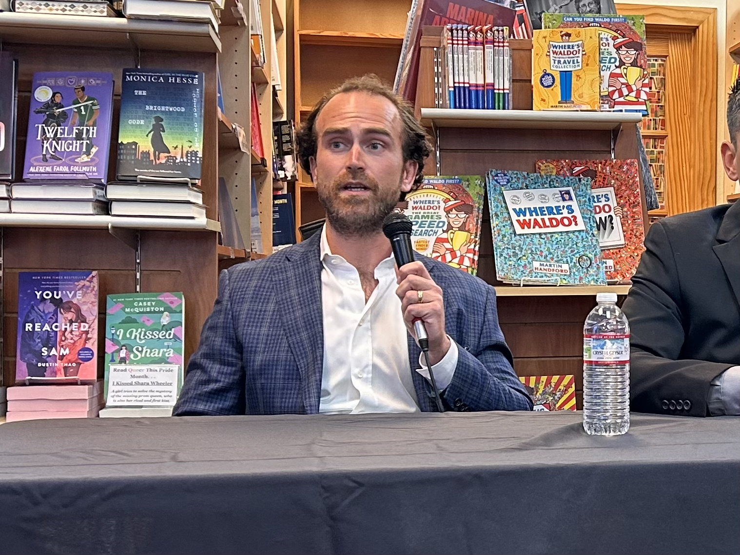 A man with curly hair and a beard, wearing a blue blazer, speaks into a microphone at a table in front of a bookshelf filled with books. A water bottle is on the table.