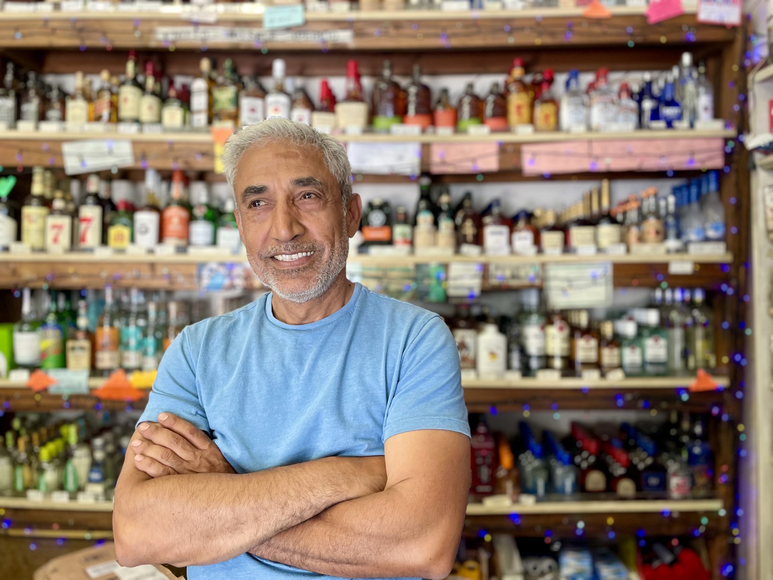 A man with folded arms stands in front of shelves filled with various bottles in a shop or store setting.