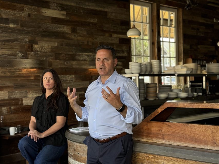 A man is speaking with gestures in a restaurant with wood-paneled walls. A woman is seated next to him, looking at him attentively, with another person in the background.