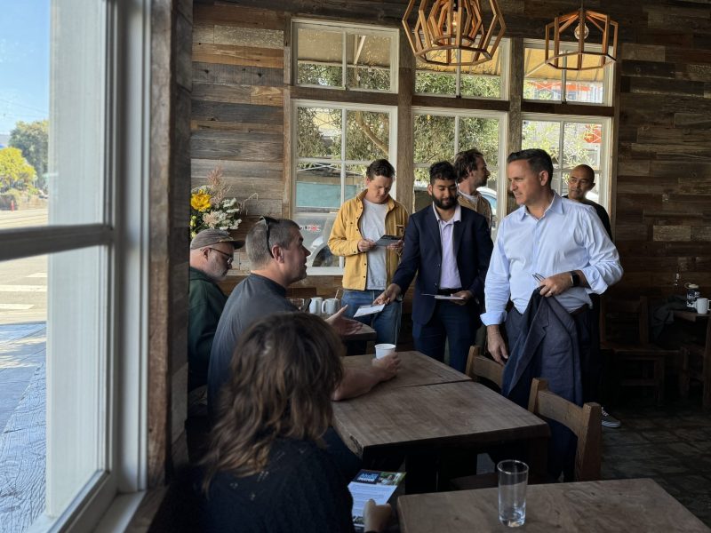 A group of people are gathered inside a wood-paneled café, engaging in conversation. Some are seated at tables while others stand, holding cups or jackets. Hanging lights illuminate the space.