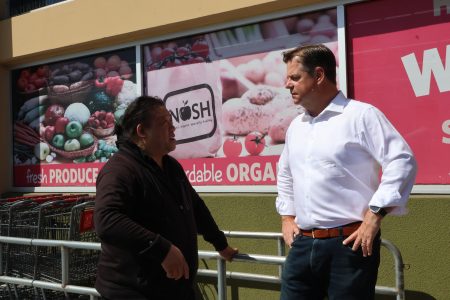 Two individuals stand and converse in front of a grocery store with "fresh PRODUCE" and "affordable ORGANIC" signs, reminiscent of Mark Farrell's community-focused projects. One person leans against a metal railing.