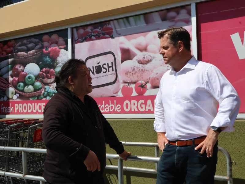 Two individuals stand and converse in front of a grocery store with "fresh PRODUCE" and "affordable ORGANIC" signs, reminiscent of Mark Farrell's community-focused projects. One person leans against a metal railing.