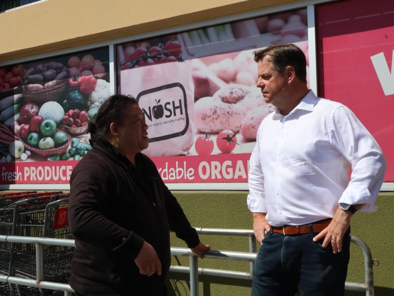 两个人站在一家杂货店前交谈，店里挂着“新鲜农产品”和“平价有机食品”的招牌，让人想起麥法恩的社区项目。一个人靠在金属栏杆上。