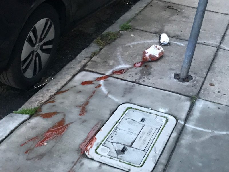 A spilled drink and food container, possibly ketchup, litter a sidewalk next to a utility cover and a parked car.