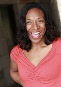 A woman with dark curly hair, wearing an orange top, stands smiling broadly inside a stone-walled structure.