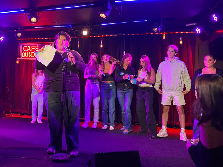 一个人站在舞台上的麦克风前，读着一张纸。旁边站着几个人，面带微笑，观察着这一切。背景上点亮了五彩灯光。