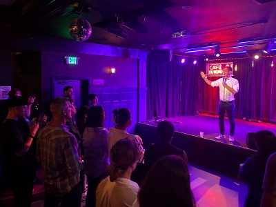 A person is performing on stage at a venue named "Cafe" with an audience watching attentively. A neon sign glows in the background.