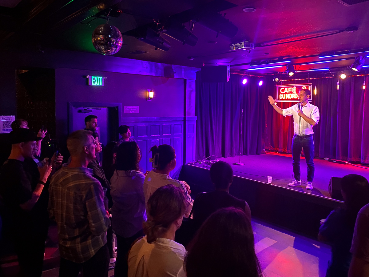 A person is performing on stage at a venue named "Cafe" with an audience watching attentively. A neon sign glows in the background.