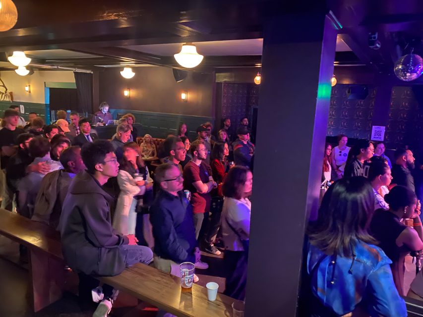 一群不同的人聚集在灯光昏暗的房间里，房间里摆放着一排排木制长椅，墙壁上贴着墙纸，挂着吊灯，专注地观看着一场表演。