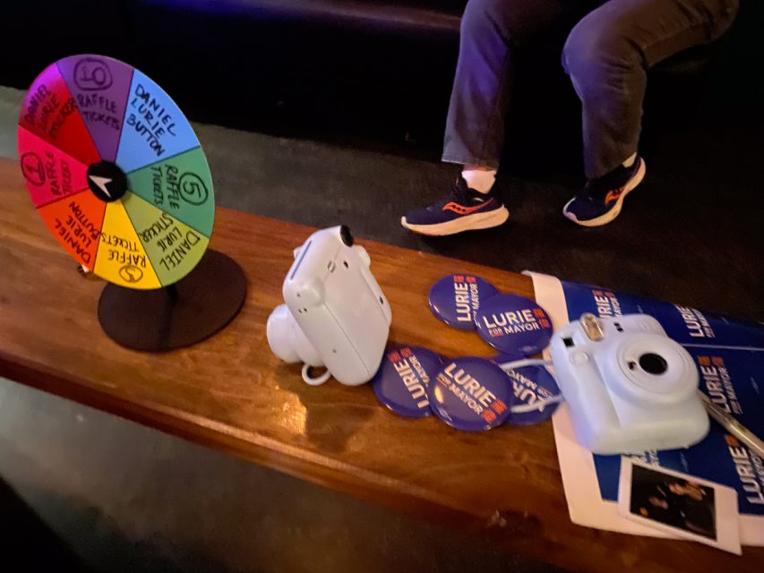 桌面上摆放着彩色奖品转盘、Instax 相机和几张“羅偉竞选市长”竞选徽章和宣传册。背景中坐着一个人。