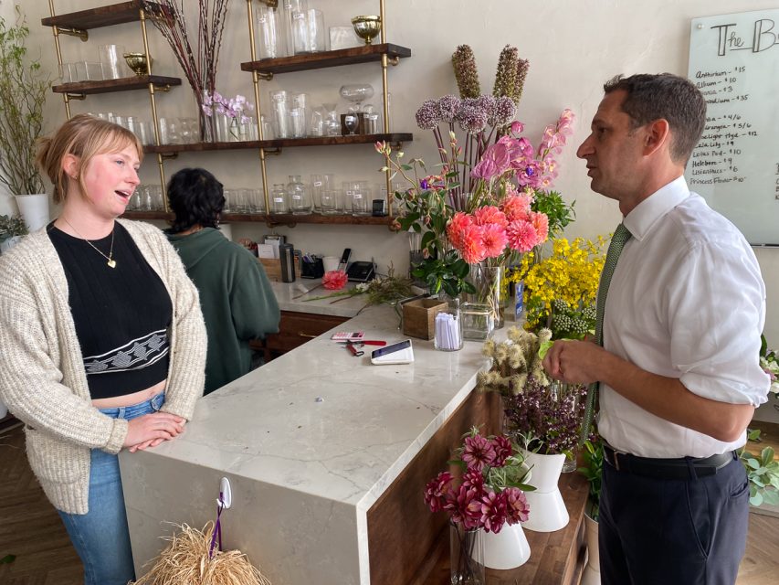 A woman in a cardigan and a man in a tie are talking across a marble counter in a flower shop. The counter displays various flowers, and shelves with jars and plants are visible in the background.