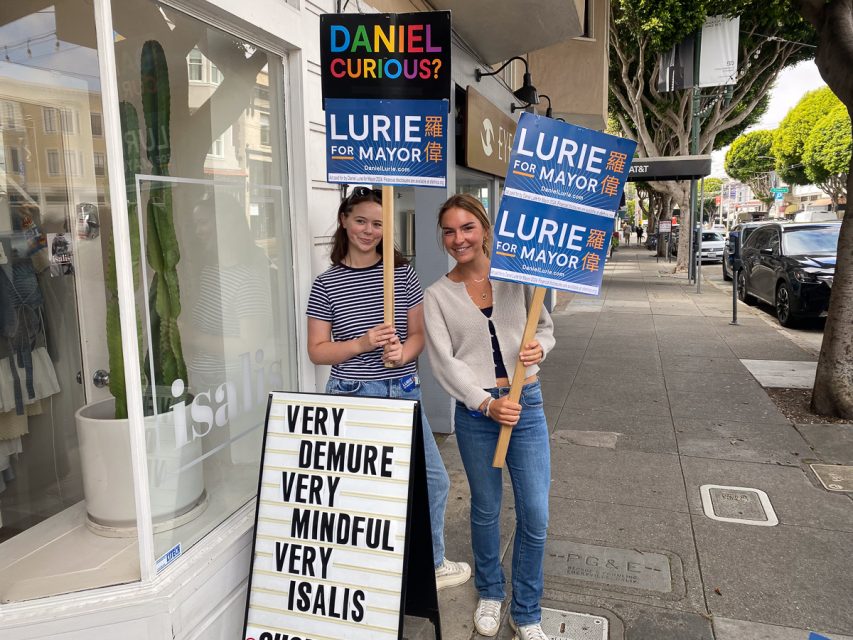 Two people standing on a sidewalk holding "Lurie for Mayor" signs. Behind them is a store with a sandwich board displaying messages like "Very Demure," "Very Mindful," and "Very Isalis.