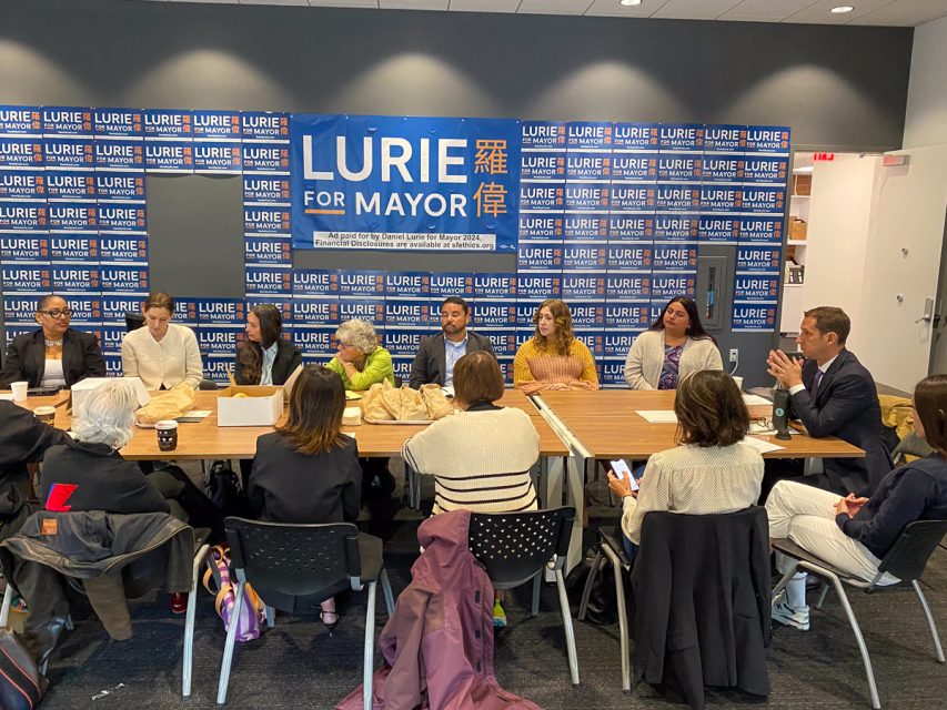 會議室裡，一群人圍坐在一張長桌旁。他們身後的競選標語寫著「羅偉競選市長」。有些人有筆記本和咖啡杯。