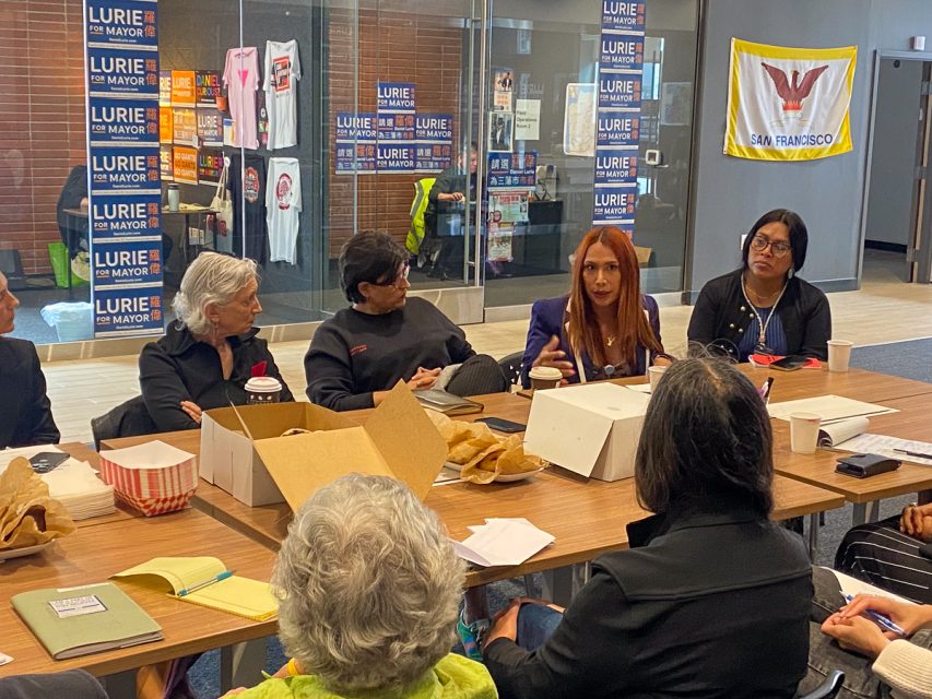 一群人圍坐在一張放著食物容器和紙張的桌子旁，進行討論。房間裡貼有競選海報，牆上掛著一面旗幟。一個人似乎正在積極發言。