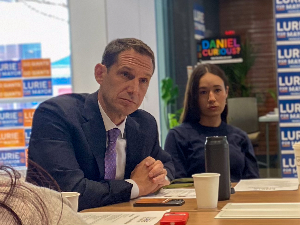 一名身穿西裝的男子和一名女子坐在桌旁，旁邊放著文件和飲料，周圍都是競選海報。