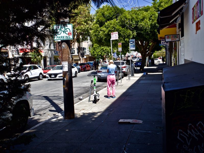 A person in pink pants walks down a city sidewalk next to a bicycle parking pole and parked electric scooters.