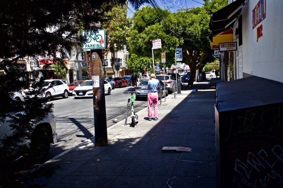 A person in pink pants walks down a city sidewalk next to a bicycle parking pole and parked electric scooters.