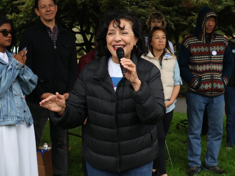 A woman wearing a black puffer jacket speaks into a microphone in an outdoor setting while several people stand behind her, listening attentively.