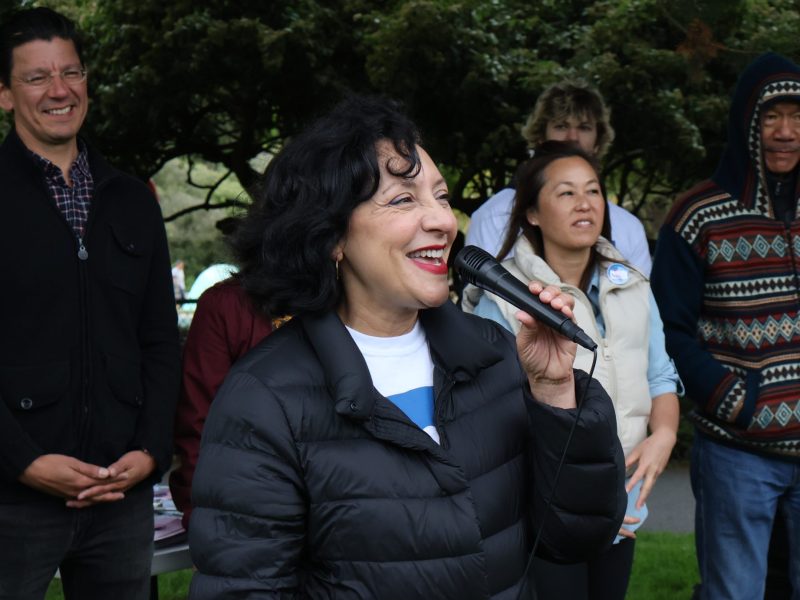 A woman in a black jacket speaks into a microphone while smiling. A group of people stand behind her, some smiling. They are outdoors with trees in the background.