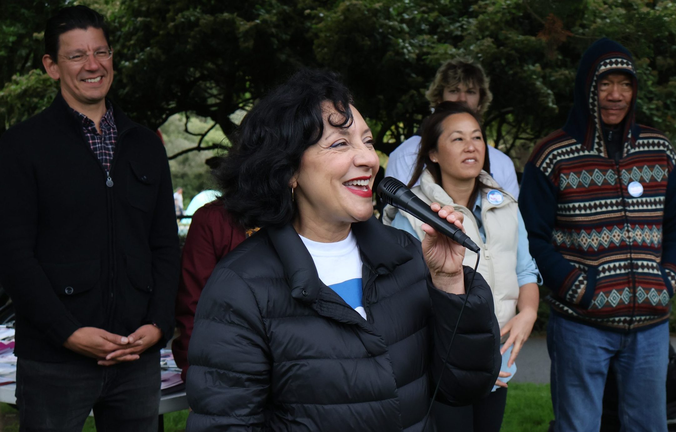 A woman in a black jacket speaks into a microphone while smiling. A group of people stand behind her, some smiling. They are outdoors with trees in the background.