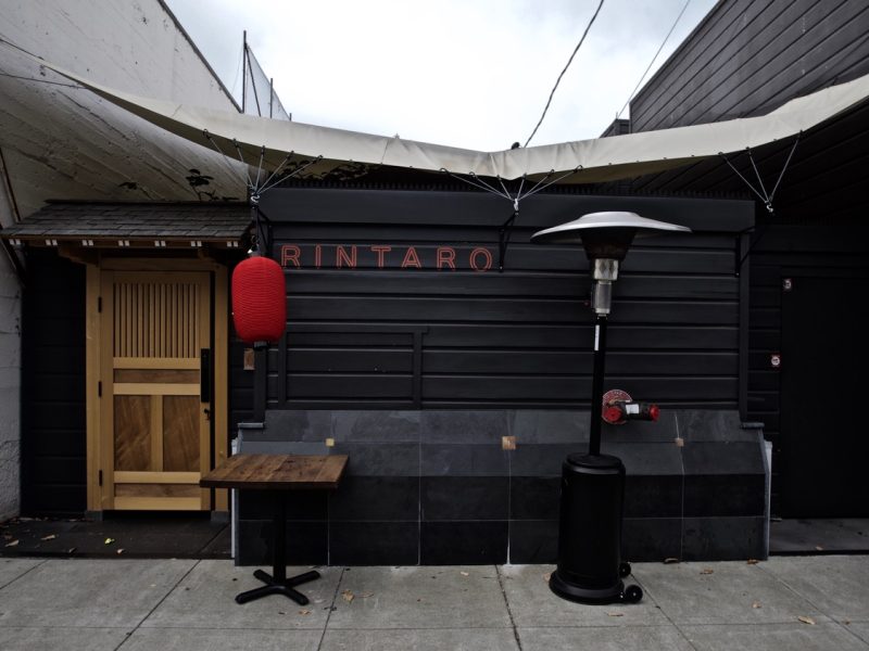 A restaurant exterior with a black facade, a wooden door, and a red lantern. The name "Rintaro" is displayed above. A patio heater and a small wooden table are placed outside.