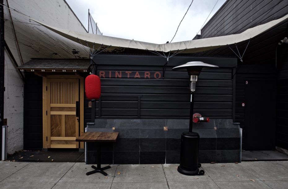 A restaurant exterior with a black facade, a wooden door, and a red lantern. The name "Rintaro" is displayed above. A patio heater and a small wooden table are placed outside.