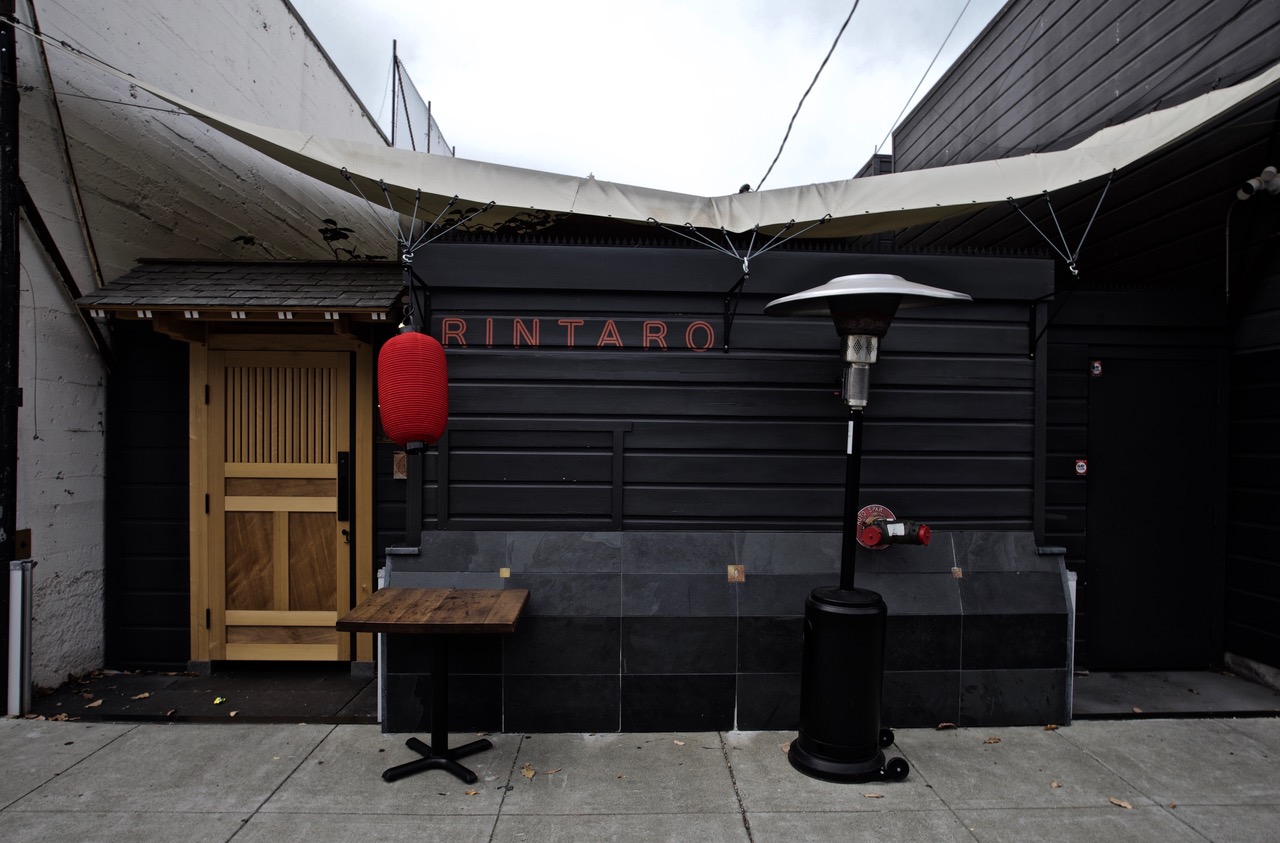 A restaurant exterior with a black facade, a wooden door, and a red lantern. The name "Rintaro" is displayed above. A patio heater and a small wooden table are placed outside.