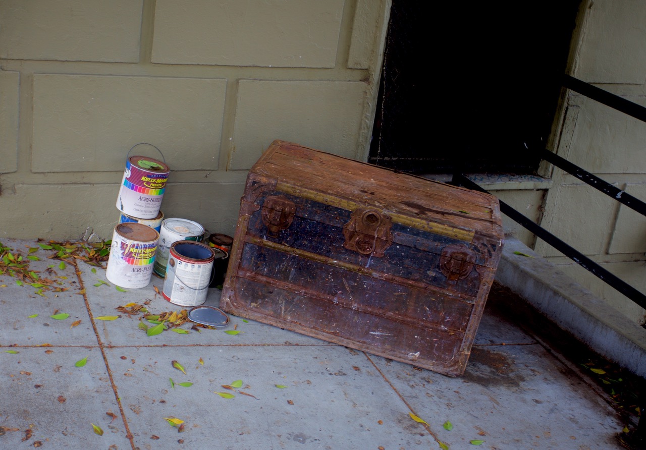 Old wooden chest and several paint cans with their lids open placed on the ground next to a building wall.
