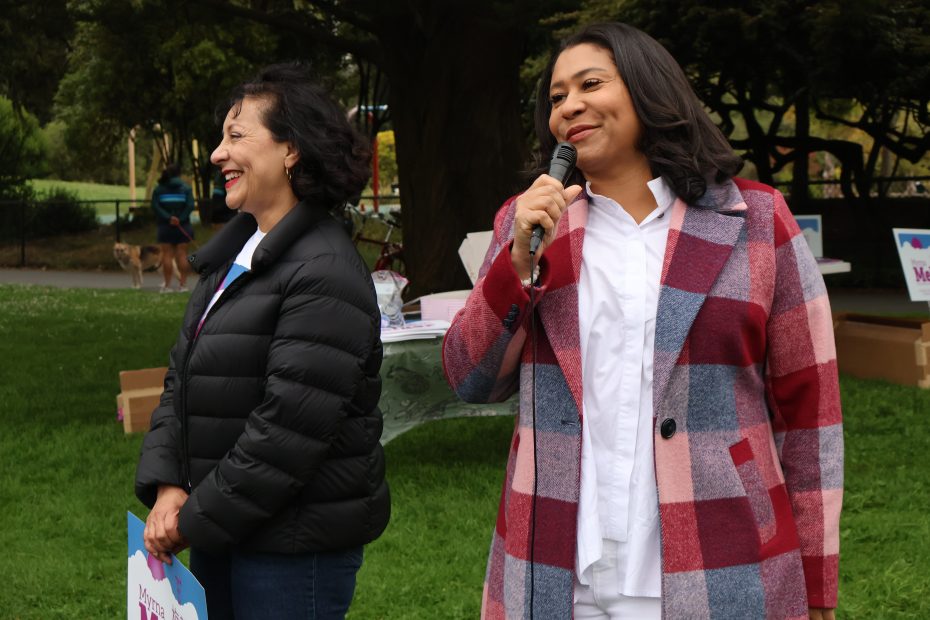 兩位女士站在戶外。一位正在對著麥克風講話，另一位在微笑。背景可見野餐桌和樹木。