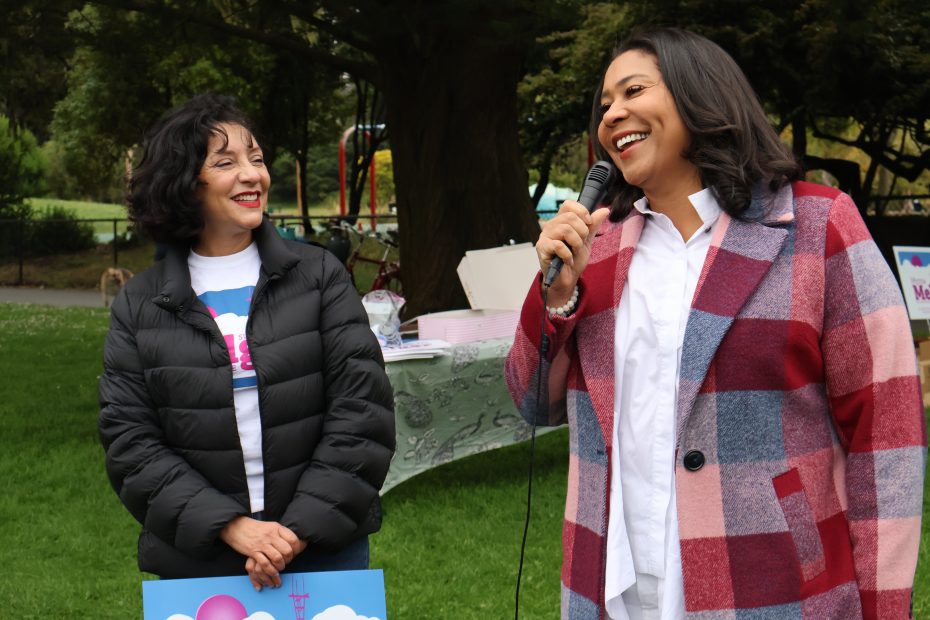 Two women are standing outdoors. One is holding a microphone and speaking, smiling, while the other woman is looking at her, also smiling. There are trees and a table behind them.