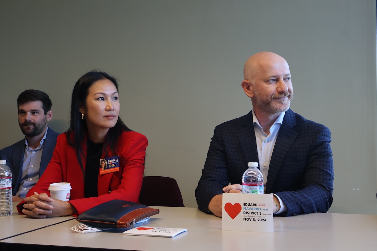 三個人坐在一張桌子旁，桌上放着文件、水瓶和一個小牌子。中間的女人穿着紅色夾克，手裡拿着一個盃子。右邊的男子拿着一個平闆電腦，上麵顯示着 "第 3 區 "的文字。