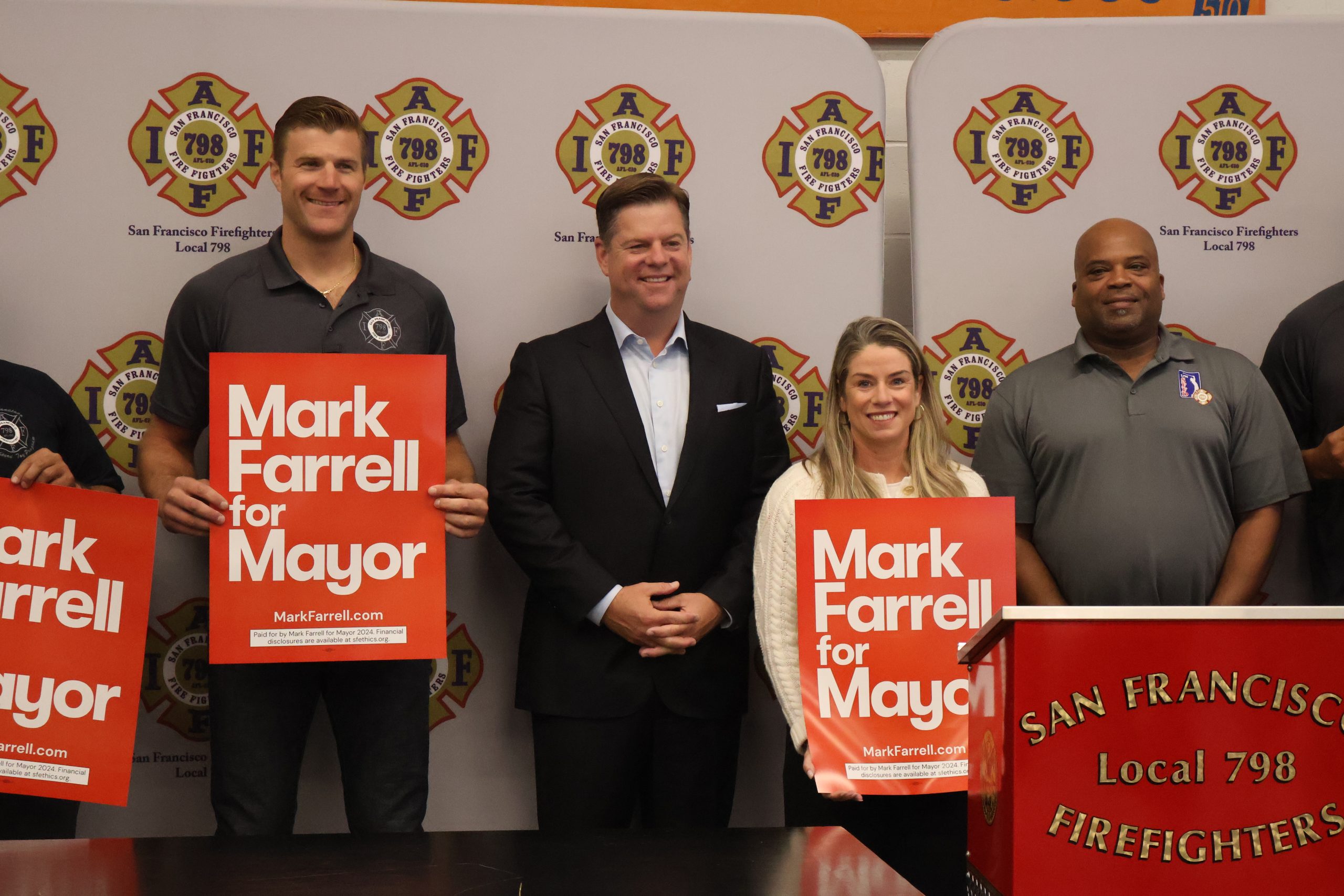 Four individuals stand together, with two holding "Mark Farrell for Mayor" signs. A podium with "San Francisco Firefighters Local 798" is in the foreground, and a backdrop features the same organization's logo.