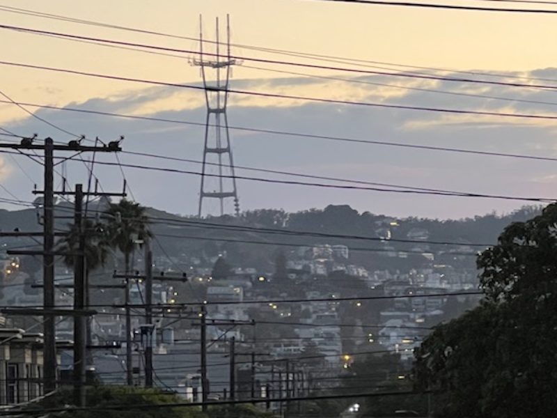 Distant view of a cityscape at dusk with power lines in the foreground and a tall transmission tower on a hill in the background.