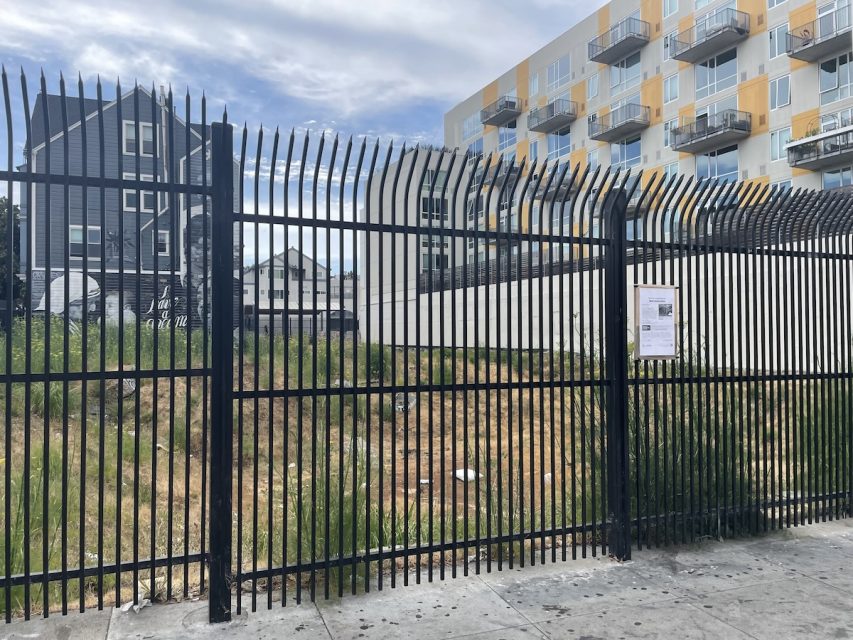 Empty fenced lot with overgrown grass and weeds in an urban area, adjacent to residential buildings. A notice is posted on the fence.