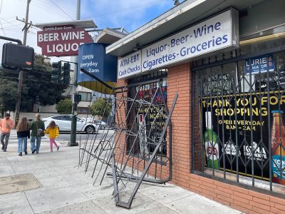 A corner store with signs advertising liquor, beer, wine, cigarettes, and groceries. There are bars protecting the windows and a metal gate partially open. People are walking on the sidewalk nearby.