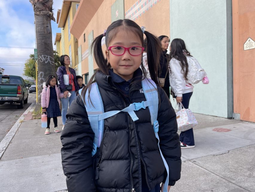 A young girl with pink glasses and pigtails stands wearing a black puffer jacket and light blue backpack. Other children and adults are in the background near a colorful building.