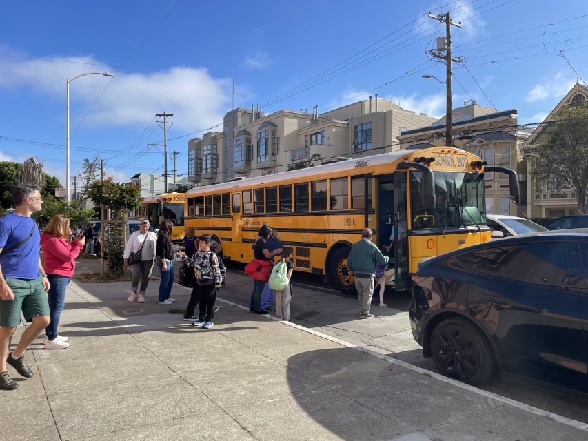 A group of people are seen boarding and standing near a yellow school bus parked on a residential street under a clear blue sky.