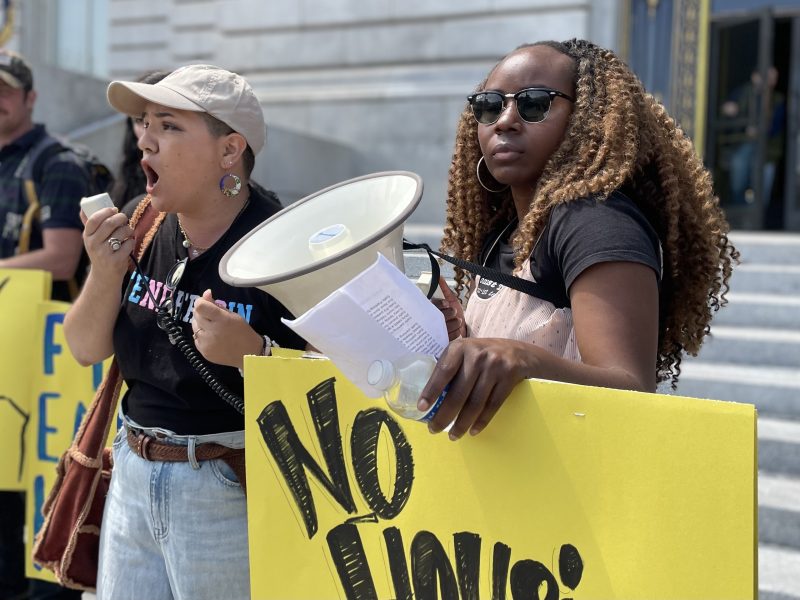 Two individuals at a protest, one holding a megaphone and the other holding a sign with "No" visible. Both wear casual clothing and stand in front of a building with steps.