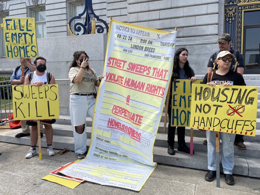 Protesters holding signs outside a building. Signs read "FINE EMPTY HOMES," "SWEEPS KILL," "STREET SWEEPS THAT VIOLATE HUMAN RIGHTS & PERPETUATE HOMELESSNESS," and "HOUSING NOT HANDCUFFS.