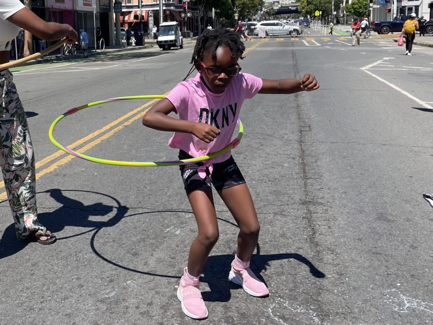 A child in pink attire and sunglasses hula hoops on an urban street, with another person standing nearby.