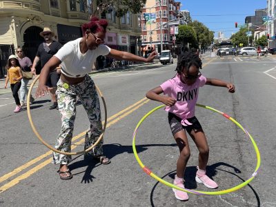 Two people hula hoop on a sunny street with buildings and pedestrians in the background. One is an adult wearing patterned pants, and the other is a child in a pink shirt and shorts.
