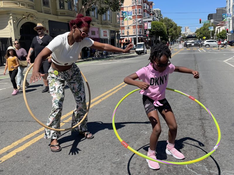 Two people hula hoop on a sunny street with buildings and pedestrians in the background. One is an adult wearing patterned pants, and the other is a child in a pink shirt and shorts.